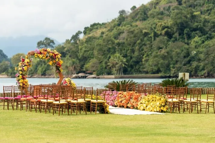 Cerimônia com arco de flores coloridas em gramado diante de uma baía