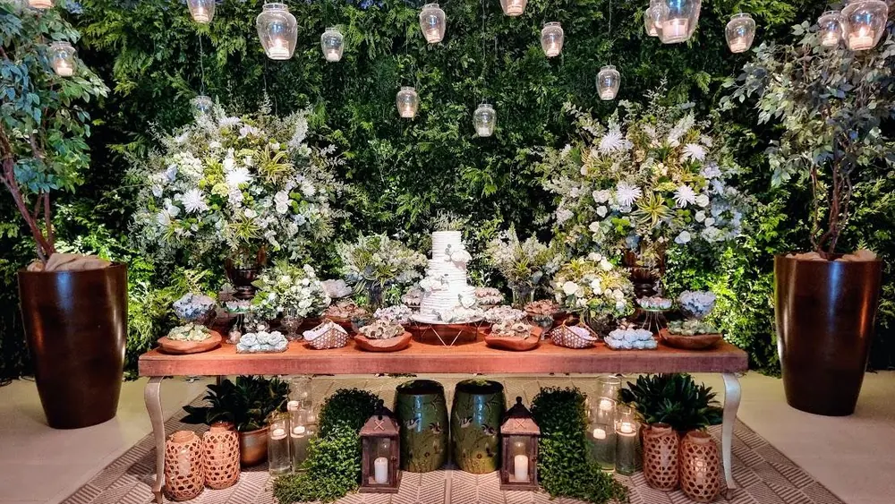 Mesa de bolo e doces diante de parede de folhagens com lanternas suspensas